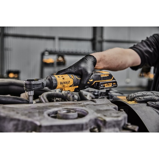 A person wearing black gloves uses a DEWALT cordless ratchet tool on an engine component in a workshop.