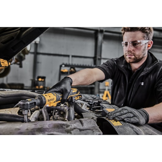 A person wearing black gloves uses a DEWALT DCF513N cordless ratchet tool to work on a car engine in a workshop.