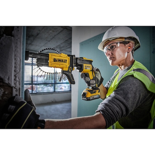 A construction worker wearing a safety helmet and vest uses a DEWALT DCF620E1K cordless drywall screw gun to fasten drywall sheets. The tool features an attached screw magazine for quick and efficient installation.