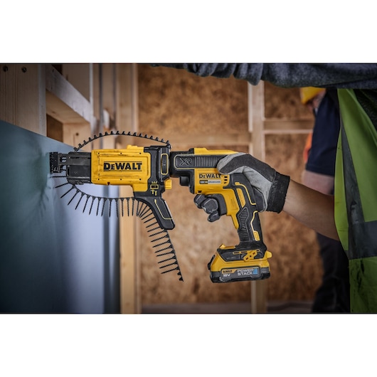 A person wearing gloves uses a DEWALT cordless screw gun with an auto-feed attachment to fasten screws into a wall panel during construction.