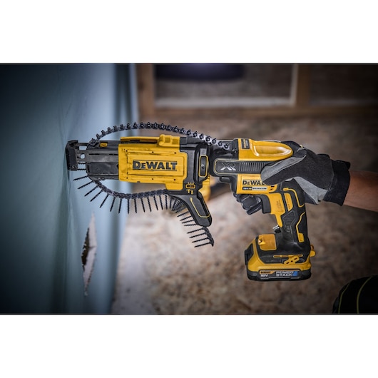 A DEWALT cordless drywall screw gun (DCF620E1K) being used to fasten screws into a wall. The tool is yellow and black, held by a gloved hand, with a strip of collated screws loaded in the gun.