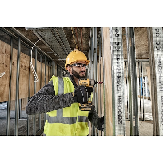 Worker using DEWALT DCF787N drill on metal studs at construction site, wearing safety gear and yellow helmet.