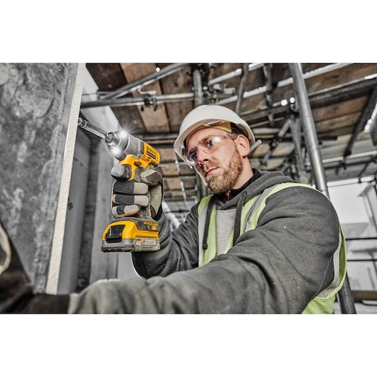 A worker wearing safety gloves, a high-visibility vest, and a helmet is using a yellow DEWALT cordless impact driver (model DCF840E1T) to drill into a concrete wall at a construction site.