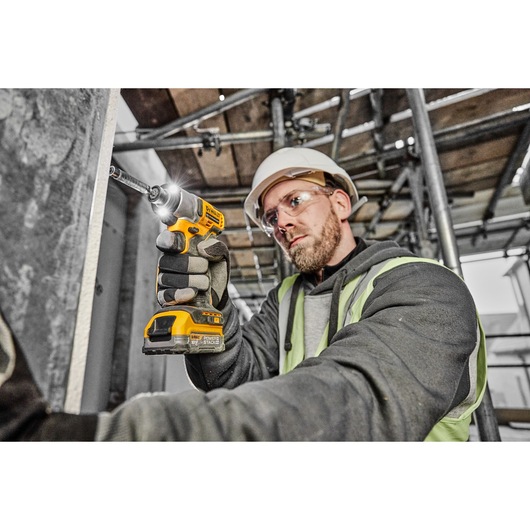 A person wearing safety gear uses a yellow DEWALT cordless drill to fasten a screw into a metal surface on a construction site.