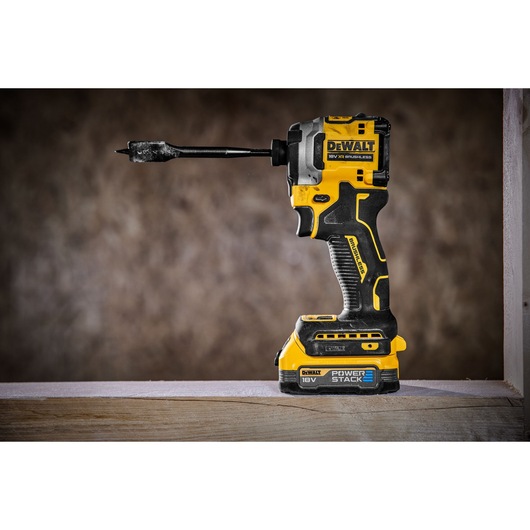 A DEWALT cordless 18V brushless impact driver with a drill bit attached and a POWERSTACK battery, standing upright on a wooden surface.