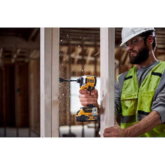 A person wearing a safety helmet and a high-visibility vest is using a DEWALT cordless drill to drill into a wooden frame at a construction site. The person's face is blurred for privacy.