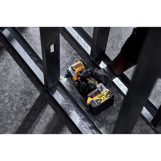 A person wearing a work glove uses a DEWALT DCF850NT cordless impact driver to fasten a screw into a metal frame on a concrete floor.