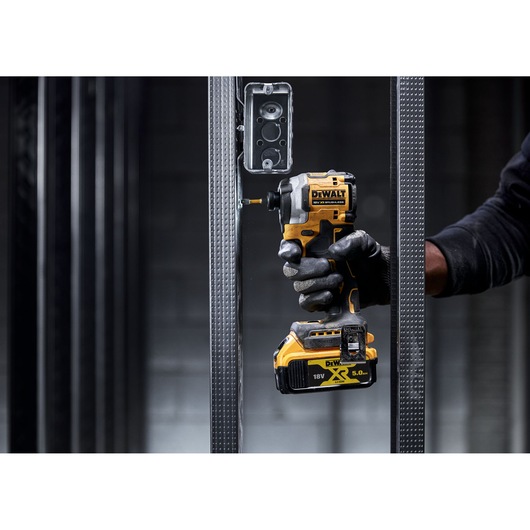 A person wearing a glove uses a DEWALT 18V XR cordless impact driver to install a metal electrical box onto a vertical metal stud frame.