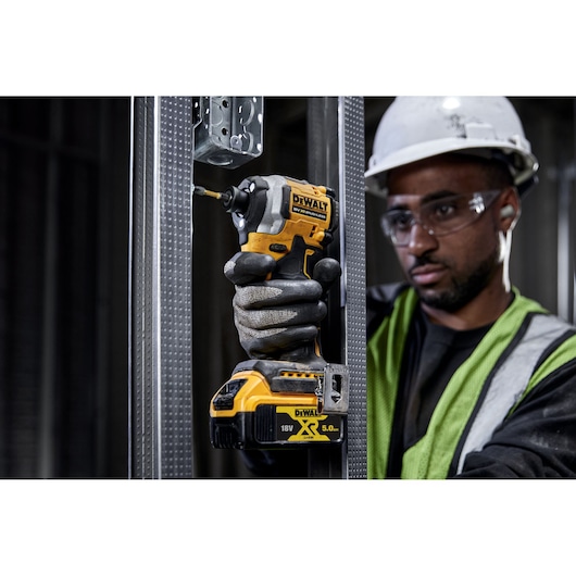 A person wearing protective gloves and a safety helmet is using a DEWALT DCF850NT 18V XR impact driver to secure a screw into a metal frame at a construction site. The person's face is obscured.