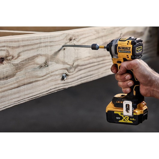 A hand holding a DEWALT 18V XR brushless impact driver, drilling a screw into a piece of light-colored wood.