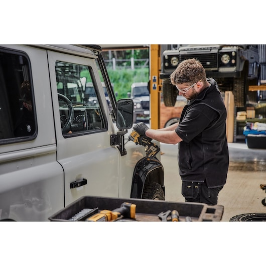 A person using a DEWALT power tool to work on the exterior of a white vehicle in a workshop environment. The person's face is blurred and they are wearing black clothing and gloves. Various tools are visible in the foreground, and other vehicles can be seen in the background.