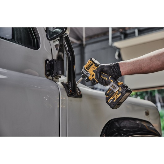 A gloved hand using a yellow and black DEWALT cordless impact driver (model DCF850NT) to fasten bolts on the exterior hinge of a vehicle door.