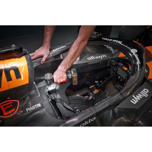 A person using a DEWALT cordless power tool (model DCF85MM1T) to work inside the cockpit of a black and orange racing car. The tool is being used near the seat and dashboard area.