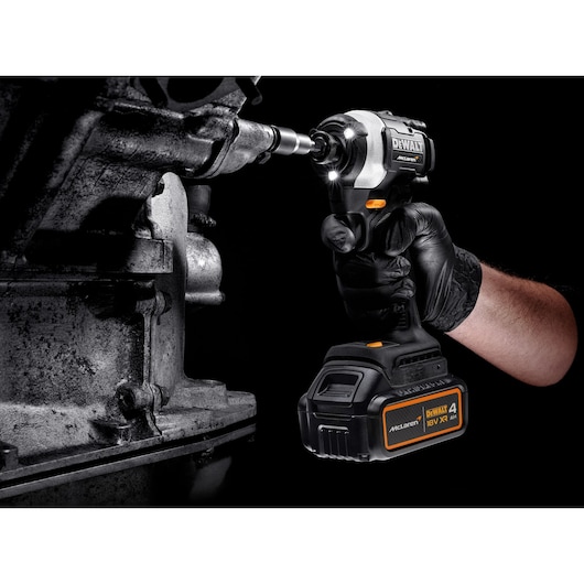 A close-up of a person wearing a black glove using a DEWALT McLaren branded cordless impact driver on a mechanical component. The tool is equipped with a 18V battery and features LED lights illuminating the work area.