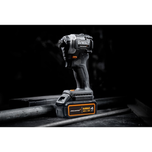A DEWALT McLaren 18V XR cordless impact driver with a 4Ah battery is shown standing on a metal surface. The tool features a black and orange color scheme.