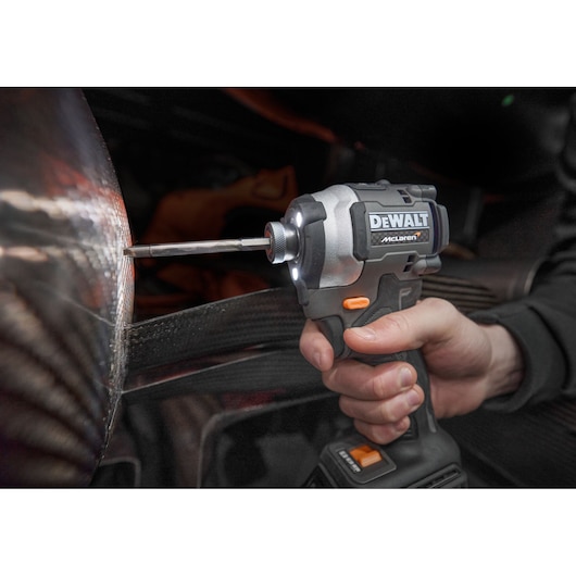 A hand holding a DEWALT cordless impact driver, model DCF85MM2T, branded with McLaren, in use against a metallic surface.