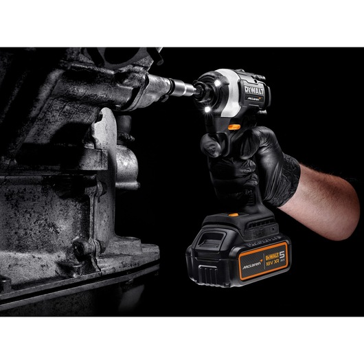 A gloved hand using a DEWALT McLaren 18V cordless impact driver with a 5Ah battery to work on a metal machine part. The tool is illuminated by built-in LED lights.