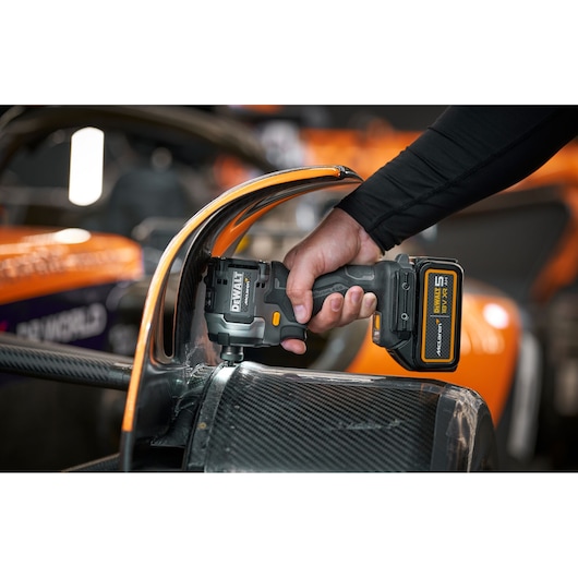 A person uses a DEWALT cordless impact wrench on part of a carbon fiber race car, with orange and black elements visible in the background.