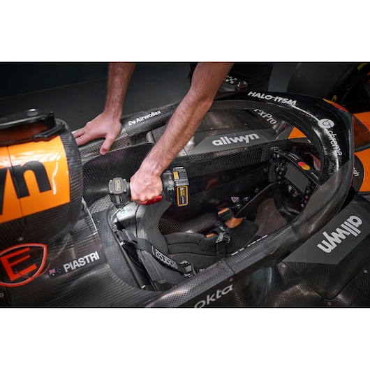 A person uses a DEWALT cordless drill on the seat area of a Formula 1 race car, focusing on the interior cockpit. The car is black and orange with various sponsor logos visible.