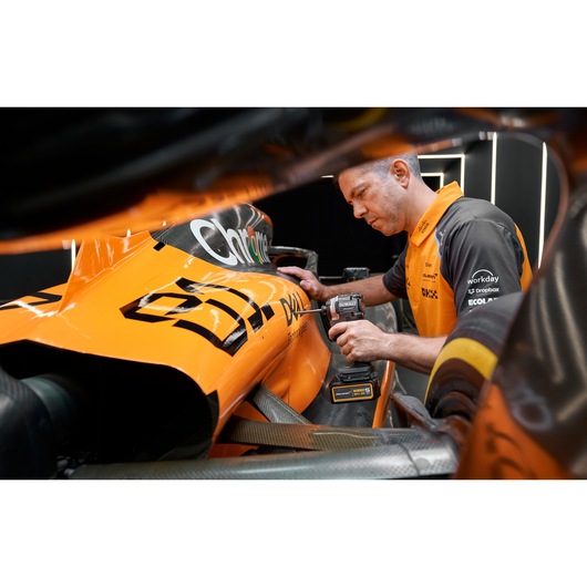 A person using a DEWALT cordless power tool to work on the side of an orange racing car in a garage or pit area. The person is wearing an orange and black shirt, and various sponsorship logos are visible on the car and clothing.