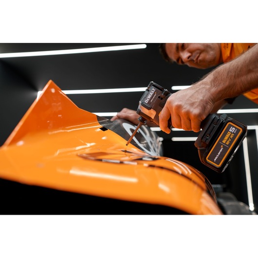 A person using a DEWALT cordless power tool to work on an orange metallic surface, likely part of a vehicle or machinery, in a well-lit room.