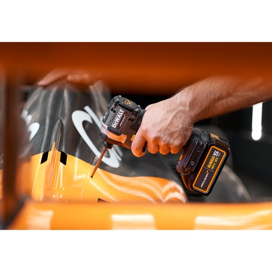A person using a DEWALT cordless impact driver to work on an orange and black surface, possibly part of a vehicle or machinery.