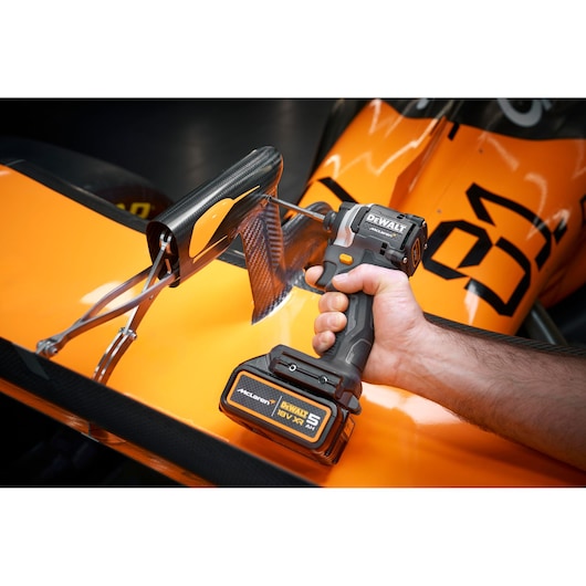 A DEWALT cordless power tool being used to work on a part of an orange race car, showing a close-up of the tool and the car's aerodynamic components.
