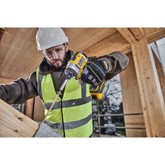 Worker uses a DEWALT DCF860E2T impact driver on wood, wearing safety gear at a construction site.