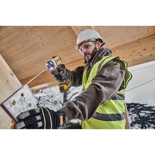 Worker uses DEWALT impact driver DCF860E2T on metal outdoors, wearing safety gear and gloves.