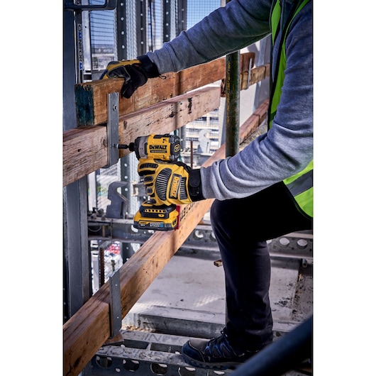 Person using a DEWALT cordless impact driver to drill into wood on a construction site, wearing gloves and safety gear.