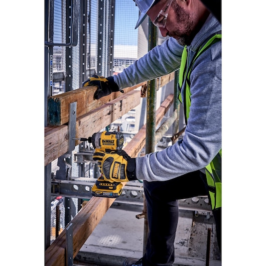 Person uses DEWALT DCF860N impact driver to drill into wood at a construction site, wearing gloves and safety gear.