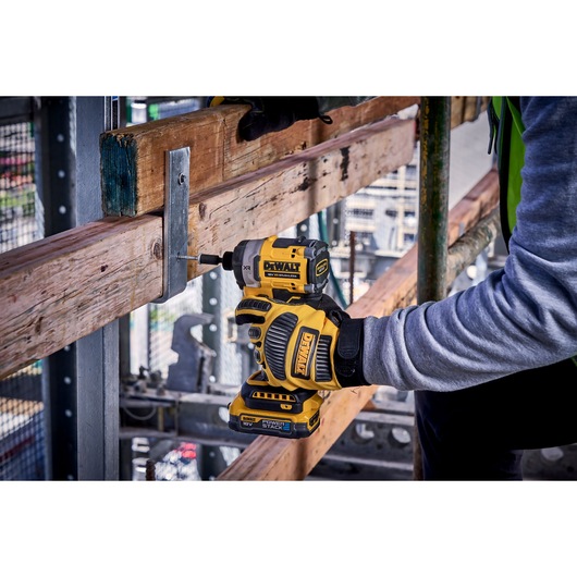 Person uses a DEWALT cordless impact driver to fasten a screw in a metal bracket on wooden beams at a construction site.
