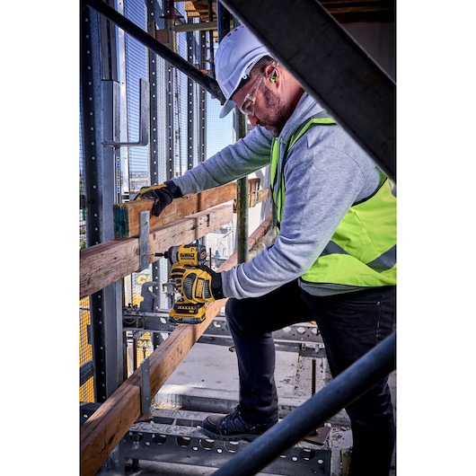 Worker uses DEWALT DCF860N impact driver on wood at construction site, wearing safety gear and high-visibility vest.