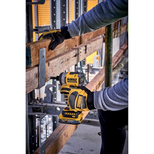 Gloved hands using a DEWALT impact driver to fasten a screw into wood at a construction site.