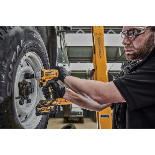 A person wearing gloves uses a DEWALT DCF891NT cordless impact wrench to loosen or tighten bolts on a vehicle wheel, inside a garage or workshop.