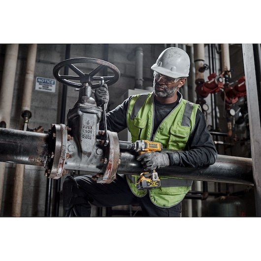 A worker wearing a safety helmet and reflective vest is using a DEWALT DCF891NT power tool on a large industrial pipe valve in a factory or plant setting. The worker's face is blurred for privacy.