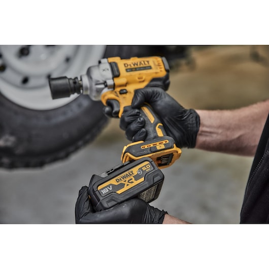 A person wearing black gloves is holding a DEWALT DCF891N cordless impact wrench and a DEWALT 18V XR 5.0Ah lithium-ion battery. The image shows the power tool and battery being prepared for use, with a vehicle tire visible in the background.