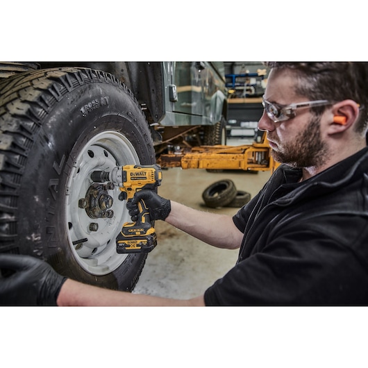 A person wearing gloves uses a DEWALT cordless impact wrench (model DCF891P2G) to work on the lug nuts of a vehicle tire in a garage setting. The person's face is blurred for privacy.
