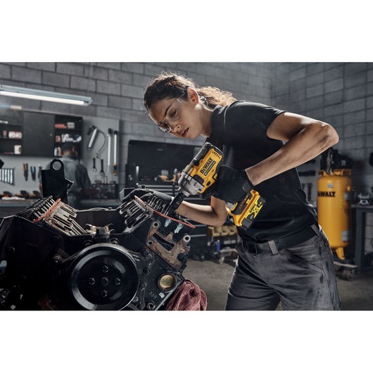 A person using a DEWALT DCF892P2T cordless impact wrench to work on a large engine component in a workshop setting. The individual is wearing gloves and has blurred facial features.
