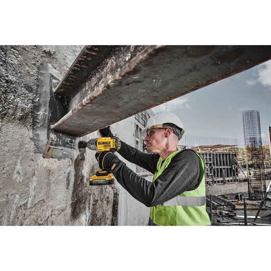 A construction worker wearing a safety helmet and high-visibility vest is using a DEWALT DCF900H2T power tool to drill into a metal beam at a construction site.