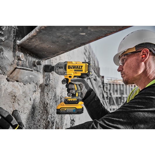 A person wearing safety gear uses a DEWALT cordless impact wrench (model DCF900H2T) with a POWERSTACK battery to tighten a bolt on a metal structure at a construction site.