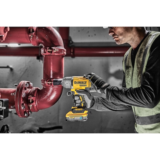 A person wearing gloves and a high-visibility vest is using a DEWALT cordless impact wrench to tighten bolts on a red industrial pipe.