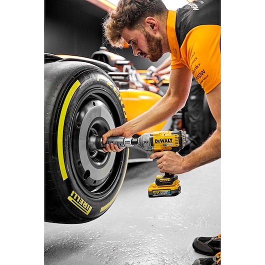A person uses a DEWALT DCF900H2T cordless impact wrench to work on the wheel of a racing car. The scene takes place in a garage, with the racing car's Pirelli tire and details of the yellow DEWALT tool clearly visible.