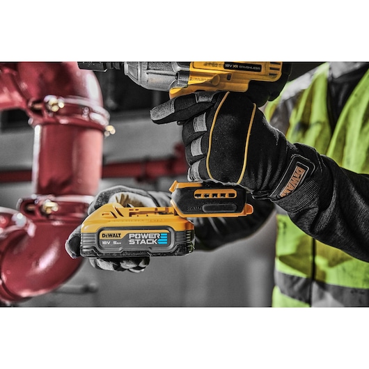 A close-up image of a DEWALT 18V XR brushless power tool being held by a person wearing black work gloves and a high-visibility vest. The tool is equipped with a DEWALT POWERSTACK 18V 5.0Ah battery. Red industrial pipes are visible in the background.