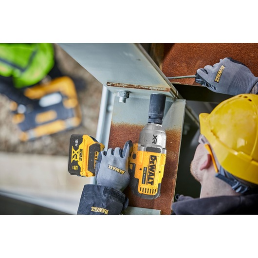 A construction worker using a DEWALT cordless impact wrench to fasten a large metal beam, wearing grey gloves and a yellow hard hat. Another person in the background is also holding DEWALT equipment.
