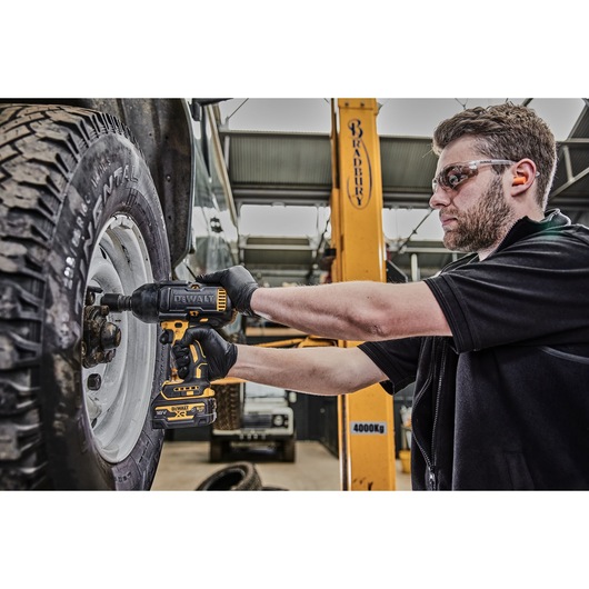 A person wearing gloves uses a DEWALT cordless impact wrench to remove or tighten lug nuts on a vehicle tire in an automotive workshop. The person's face is blurred, and there is workshop equipment and a lifted vehicle visible in the background.