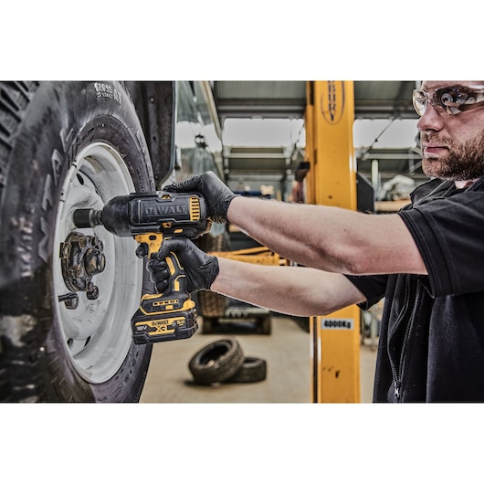 A person wearing black gloves uses a DEWALT 18V impact wrench to work on the lug nuts of a vehicle wheel in a workshop setting. Other car tires and workshop equipment are visible in the background.