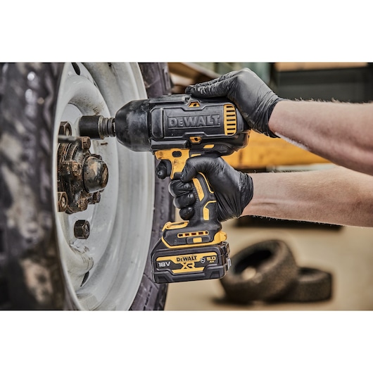 A person wearing black gloves is using a DEWALT 18V XR cordless impact wrench to loosen a lug nut on a vehicle wheel.