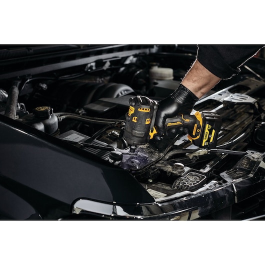 A hand wearing a black glove is using a DEWALT cordless impact wrench (model DCF901P1G) to work on the engine compartment of a car.