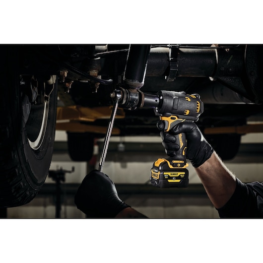 A person wearing black gloves uses a DEWALT cordless impact wrench to loosen a bolt under a vehicle. The tool is yellow and black and is being used in an automotive workshop setting.